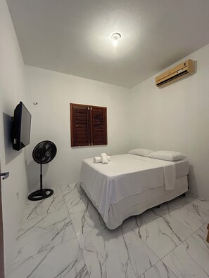 A bedroom with a bed, a wall-mounted air conditioner, a standing fan, and a television on the wall.