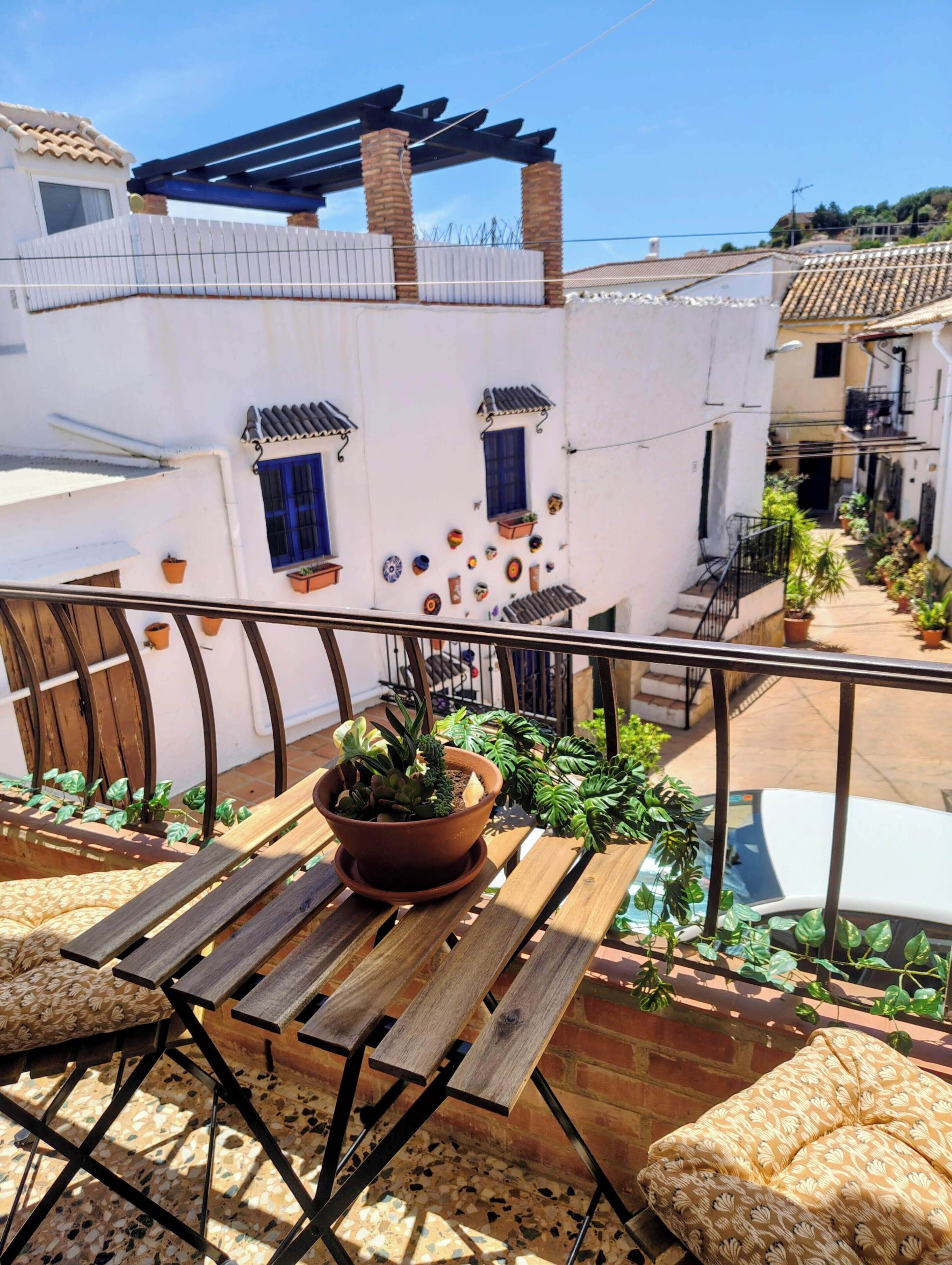 A balcony with a wooden table, potted plants, and a white building with blue windows and decorative elements.