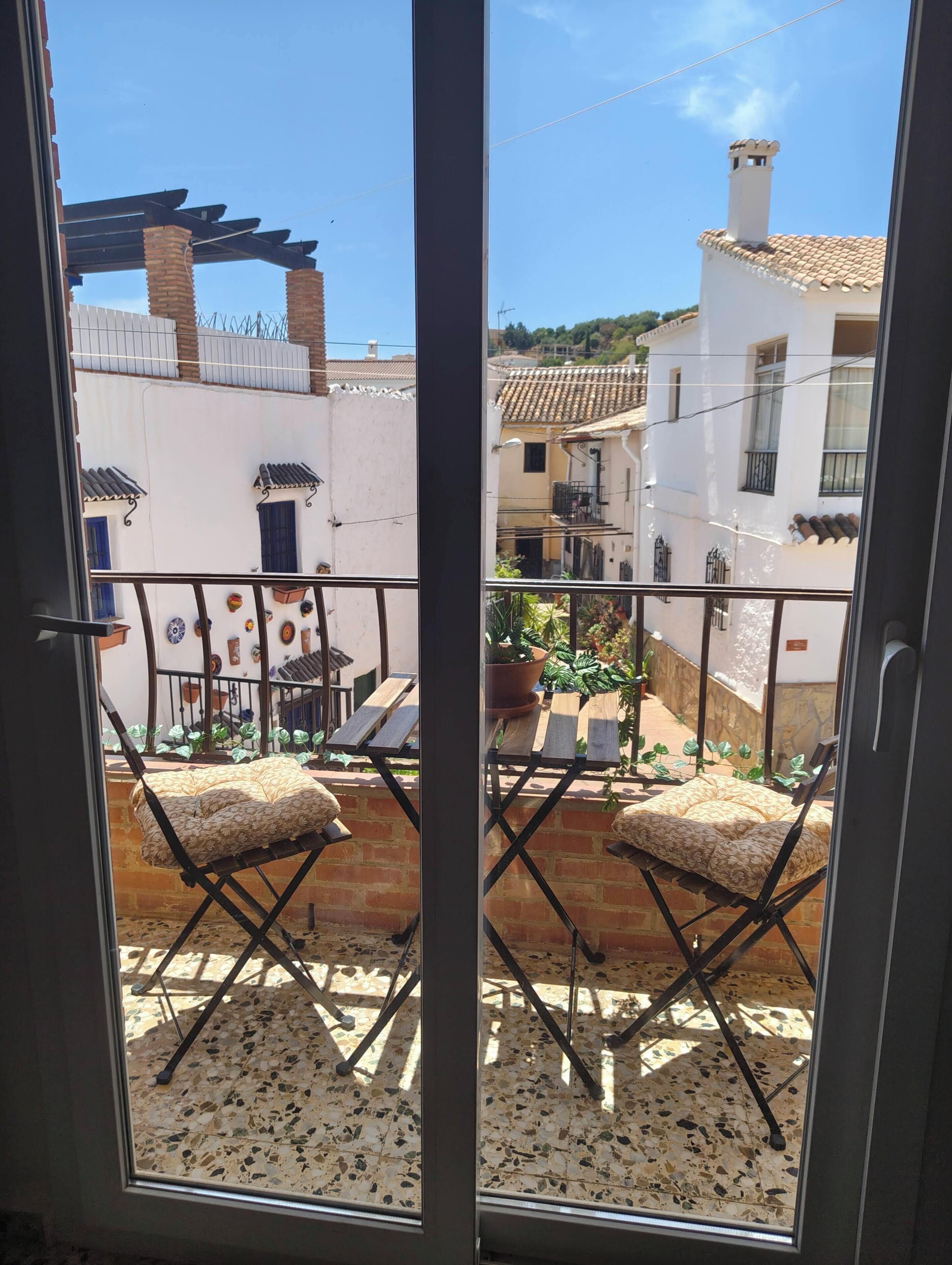 Ein Balkon mit zwei Klappstühlen und einem Blumentopf, der auf einen Innenhof mit einem weißen Gebäude und einem Ziegeldach blickt.