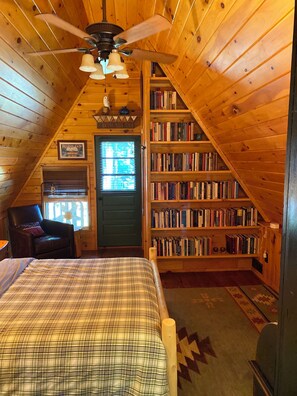 Une chambre mansardée accueillante, avec un lit, une chaise, une bibliothèque et un ventilateur de plafond.