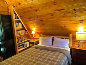 Un dormitori acollidor amb un sostre de fusta, un llit amb almoines blanques, una taula de nit amb una làmpada i una biblioteca plena de llibres.