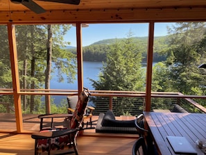 A wooden deck with a table and chairs, overlooking a lake and forest. - Charming Adirondack Retreat Overlooking Lake Luzerne (Lake Luzerne)