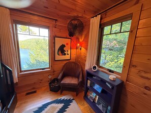 A cozy cabin room with wooden walls, a leather chair, a bookshelf, and a large window with a view of trees. - Charming Adirondack Retreat Overlooking Lake Luzerne (Lake Luzerne)