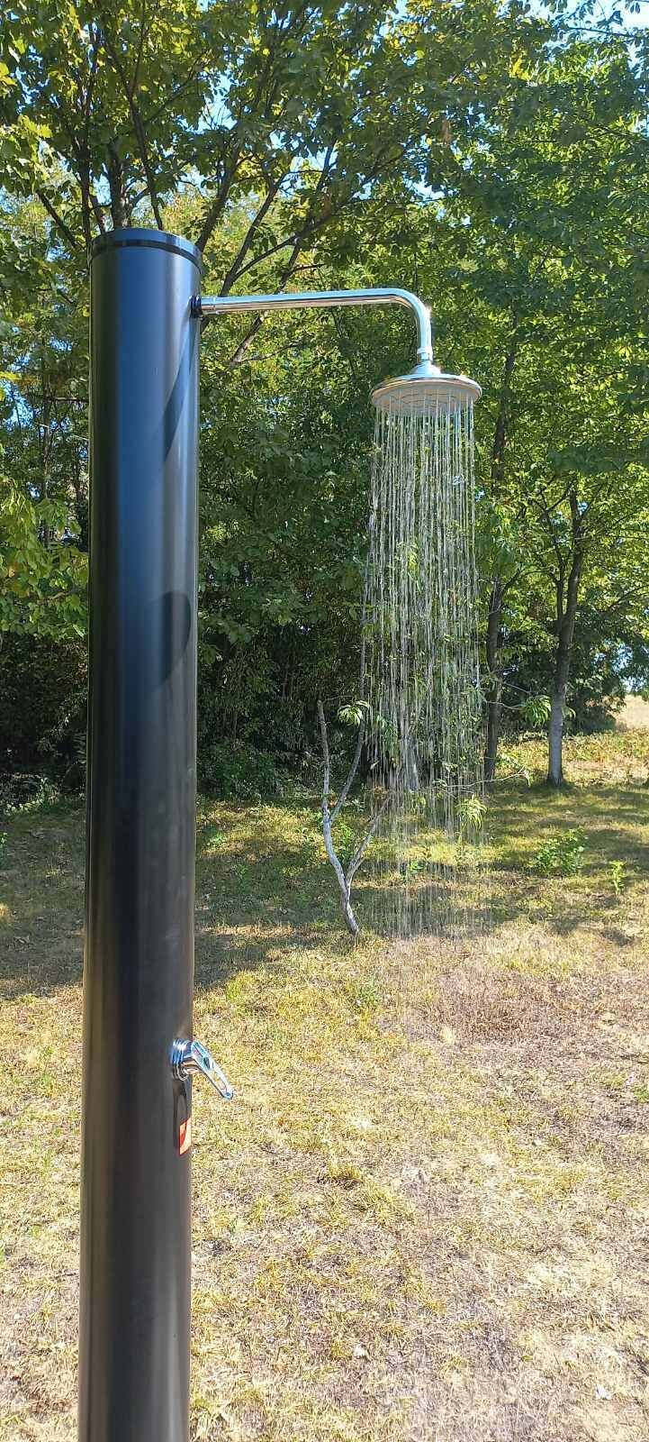 Una fuente de metal con caño y un elemento colgante, ubicada en una zona verde con árboles al fondo.