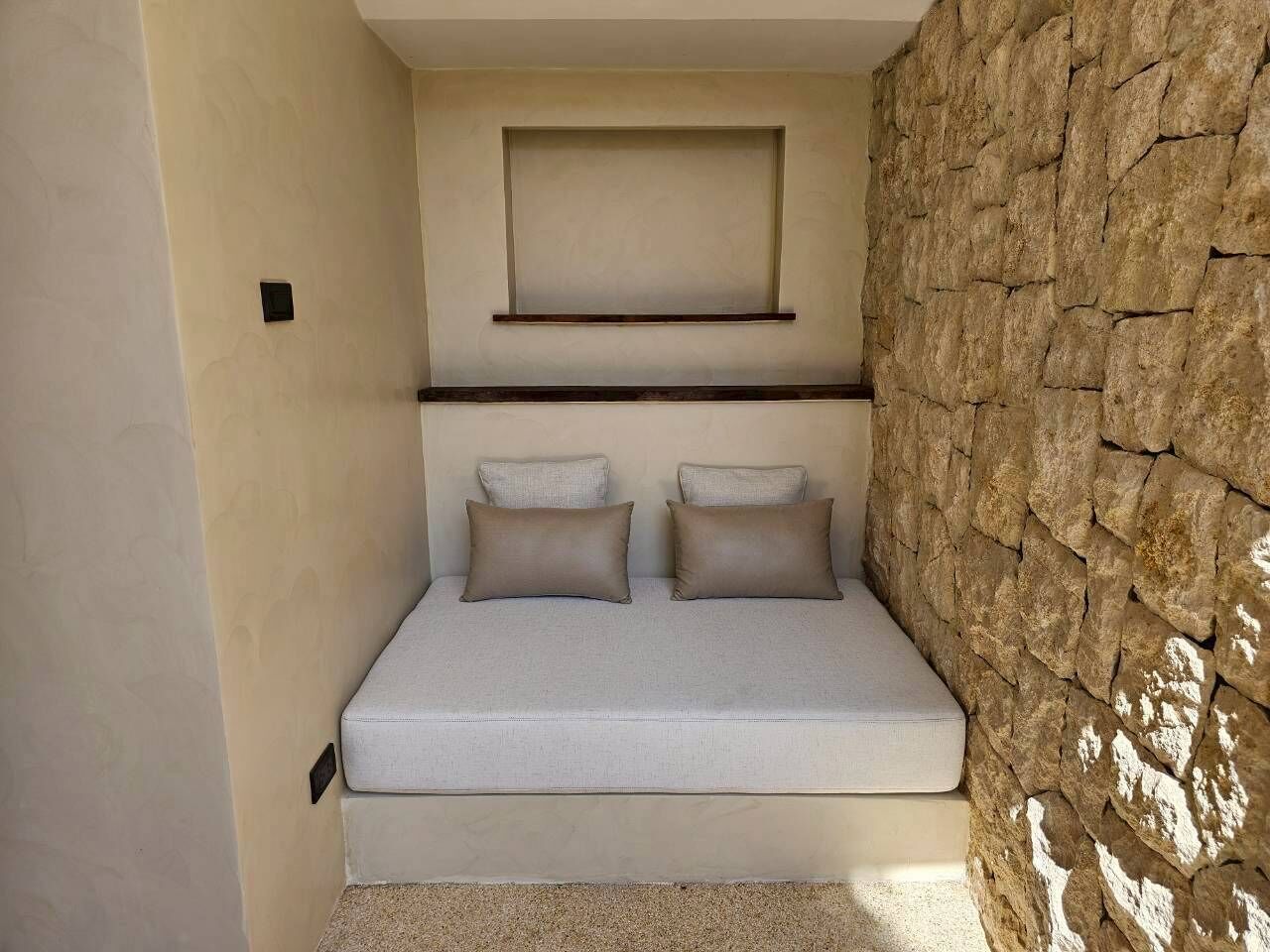 A small, cozy room with a bed, a wall-mounted shelf, and a textured stone wall.