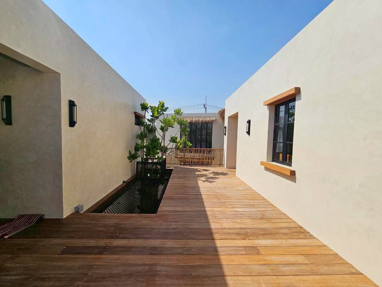 A narrow wooden pathway leading to a thatched-roof structure, flanked by plain walls with small windows.