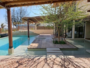 A pool area with a wooden deck, a pergola, and a modern building in the background. - Sun-kissed Retreat Beachfront villa (Cha-am)