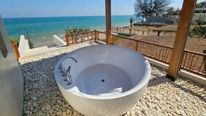 Una bañera independiente grande con un solo grifo, rodeada de guijarros blancos, bajo una pérgola de madera con vista al mar.