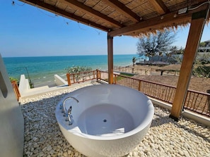 Una bañera independiente grande con un solo grifo, rodeada de guijarros blancos, bajo una pérgola de madera con vista al mar.