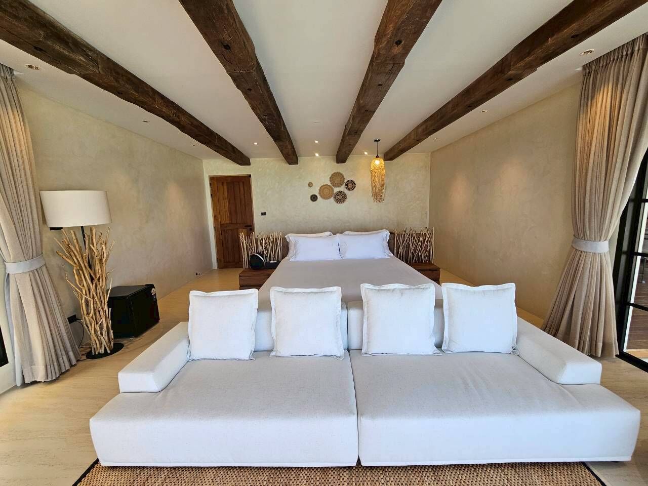 A large bed with white bedding, a sofa with white cushions, wooden beams on the ceiling, and a lamp on the left.