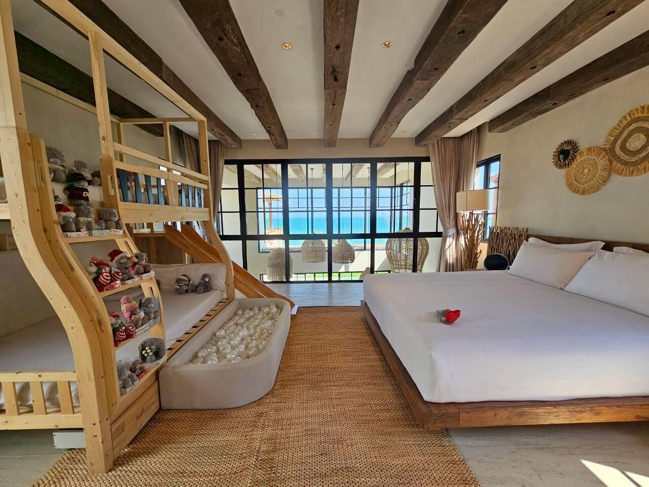 A bedroom with a large bed, a wooden crib with toys, a view of the sea, and a balcony with wicker chairs.
