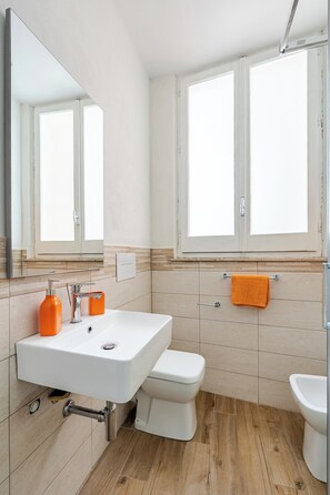 Ein Badezimmer mit einem weißen Waschbecken, einer Toilette und einem Bidet. Über dem Waschbecken hängt ein Spiegel und über der Toilette ein Fenster.
