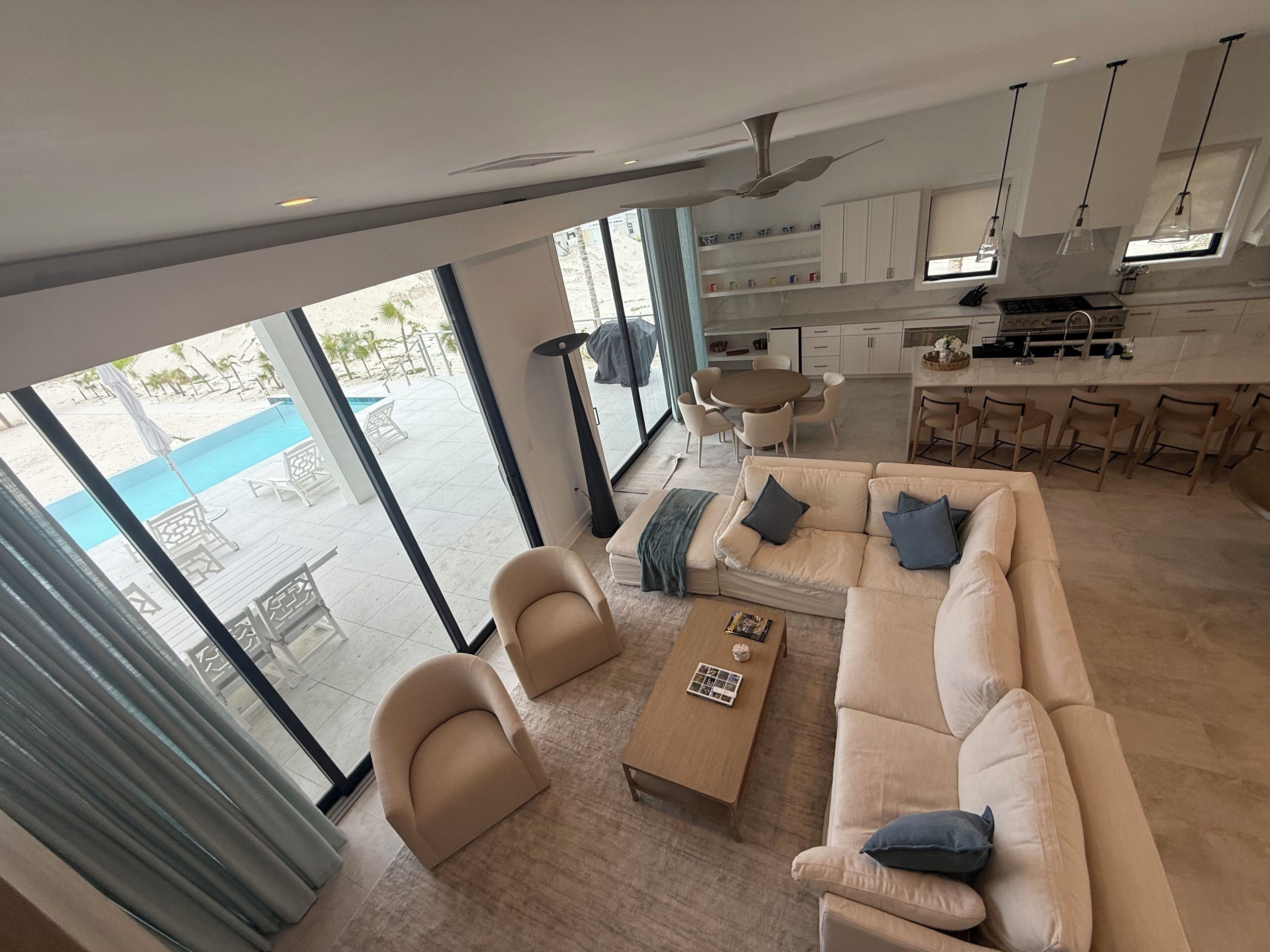 Una sala moderna con sofá, sillones y una mesa de centro. Se ve una cocina con armarios blancos y un comedor con mesa y sillas. Amplias ventanas ofrecen vistas a un área de piscina al aire libre.