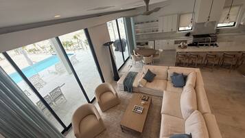 Una sala moderna con sofá, sillones y una mesa de centro. Se ve una cocina con armarios blancos y un comedor con mesa y sillas. Amplias ventanas ofrecen vistas a un área de piscina al aire libre.