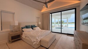 A bedroom with a large bed, a ceiling fan, a nightstand, a lamp, a bench, a dresser, and a view of a pool and beach through large glass doors. - Spectacular Beachfront Ultra Luxury Villa Stella Maris-Introductory Sale! (Sandy Point)