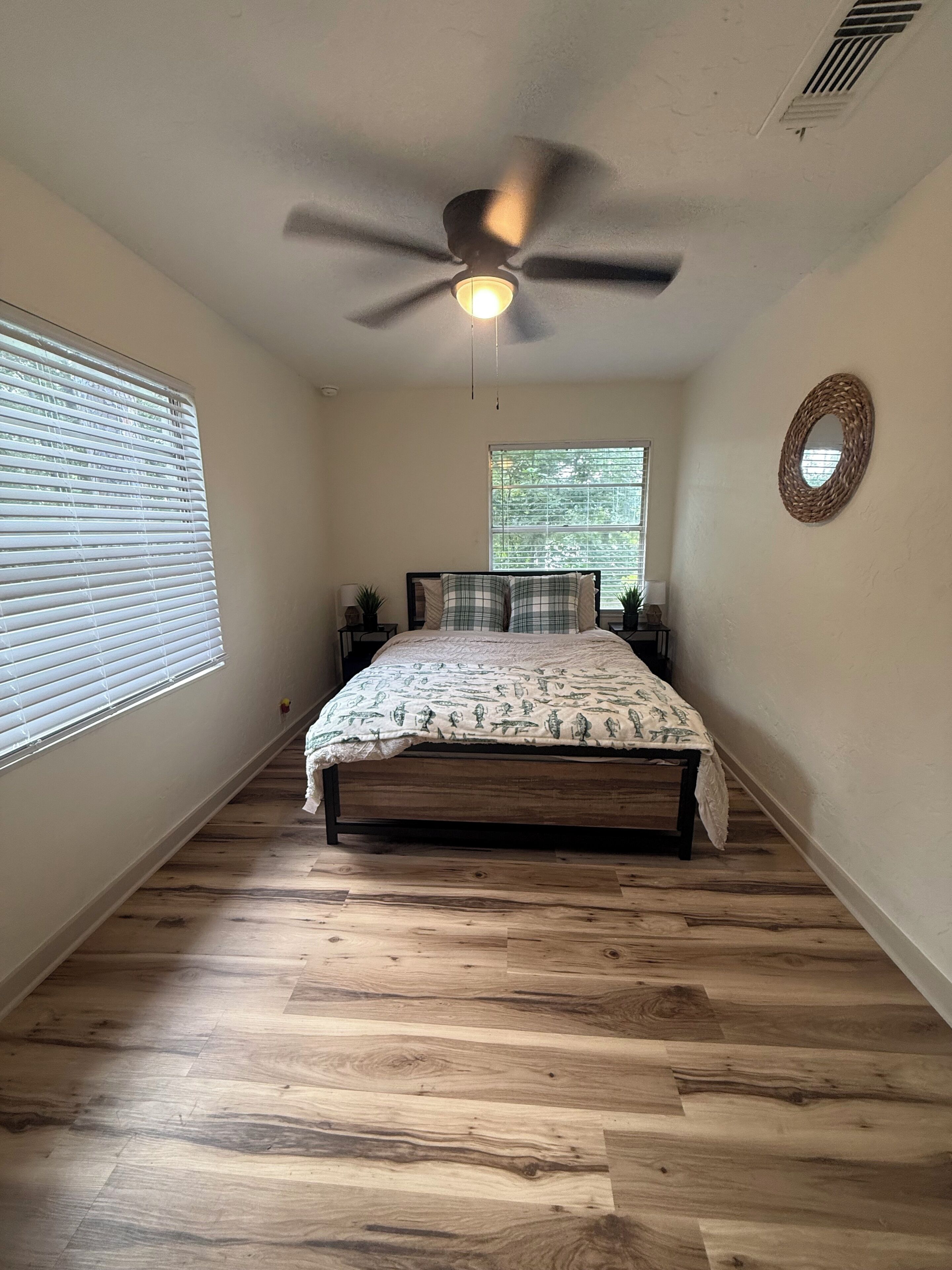 Une chambre à coucher avec un lit, un ventilateur de plafond, une fenêtre et un miroir fixé au mur.