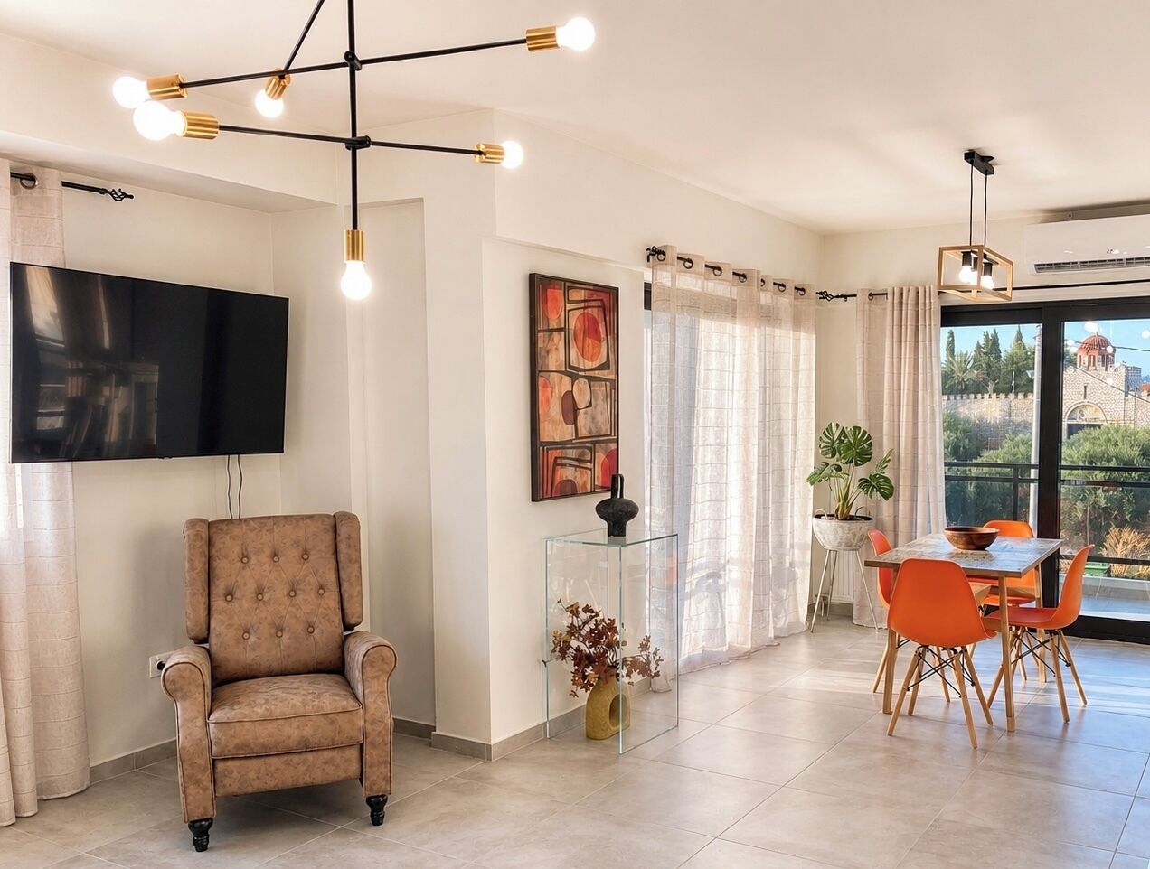 A modern living room with a flat-screen TV, a tufted armchair, a glass coffee table, and a dining area with orange chairs.