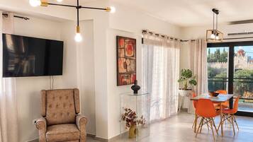 A modern living room with a flat-screen TV, a tufted armchair, a glass coffee table, and a dining area with orange chairs.