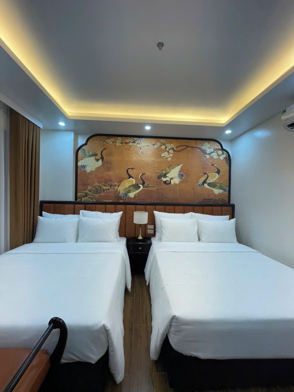 Habitación de hotel con dos camas, cabecera con mural, lámparas de noche y suelo de madera.