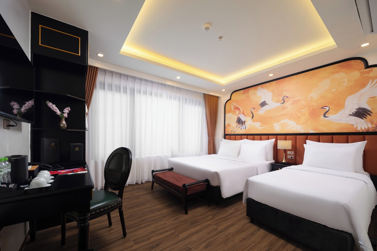 A hotel room with two beds, a desk with a chair, a small table, and a wall with a painting of birds.