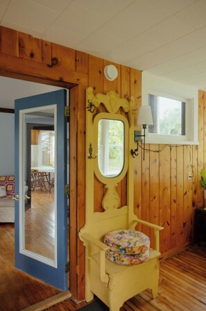 Una habitación con paredes de madera, una silla amarilla con cojín floral, un espejo y una ventana con lámpara blanca.