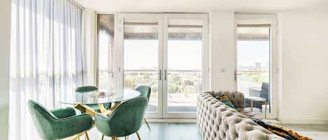 A modern living room with a glass dining table, green chairs, a tufted sofa, and a city view through large sliding doors.