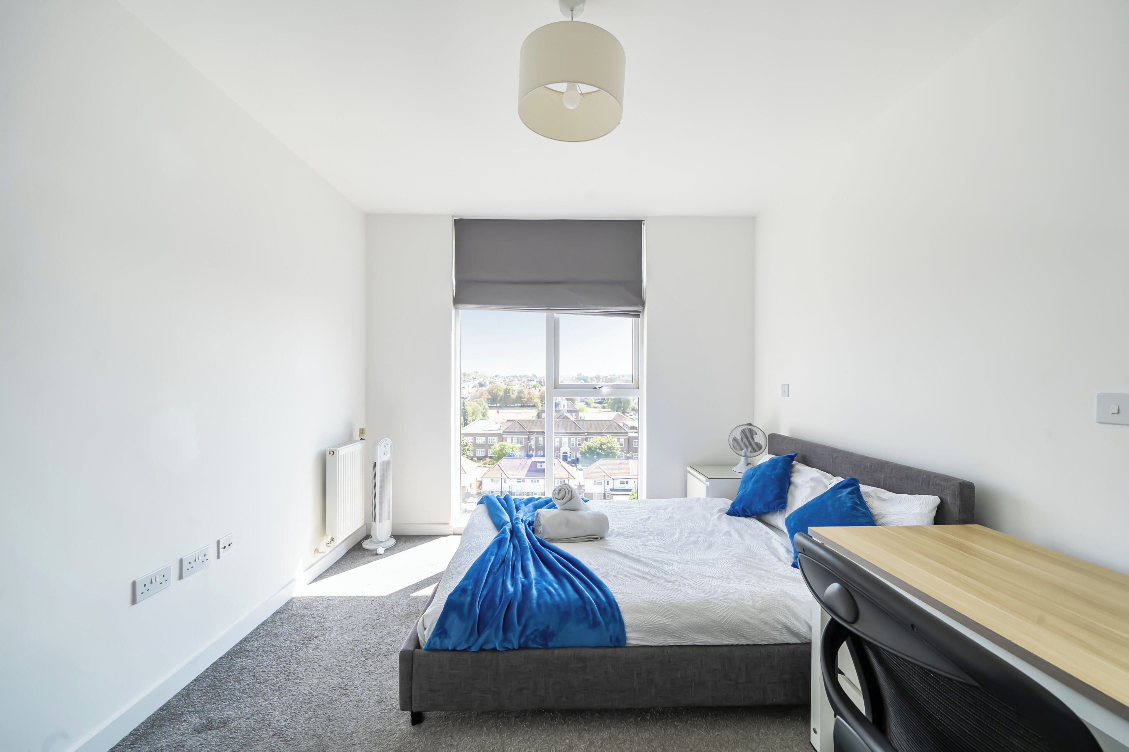 A modern bedroom with a large window, a bed with a blue blanket, a bedside table, and a radiator.