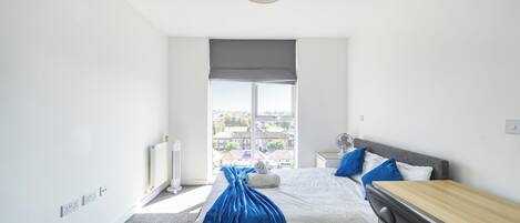 A modern bedroom with a large window, a bed with a blue blanket, a bedside table, and a radiator.