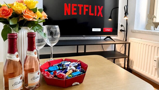 A room with a television displaying the Netflix logo, a red box of chocolates, two bottles of wine, and a vase of flowers. - N&N: Apartment - in the heart of the Old Town (Wismar)