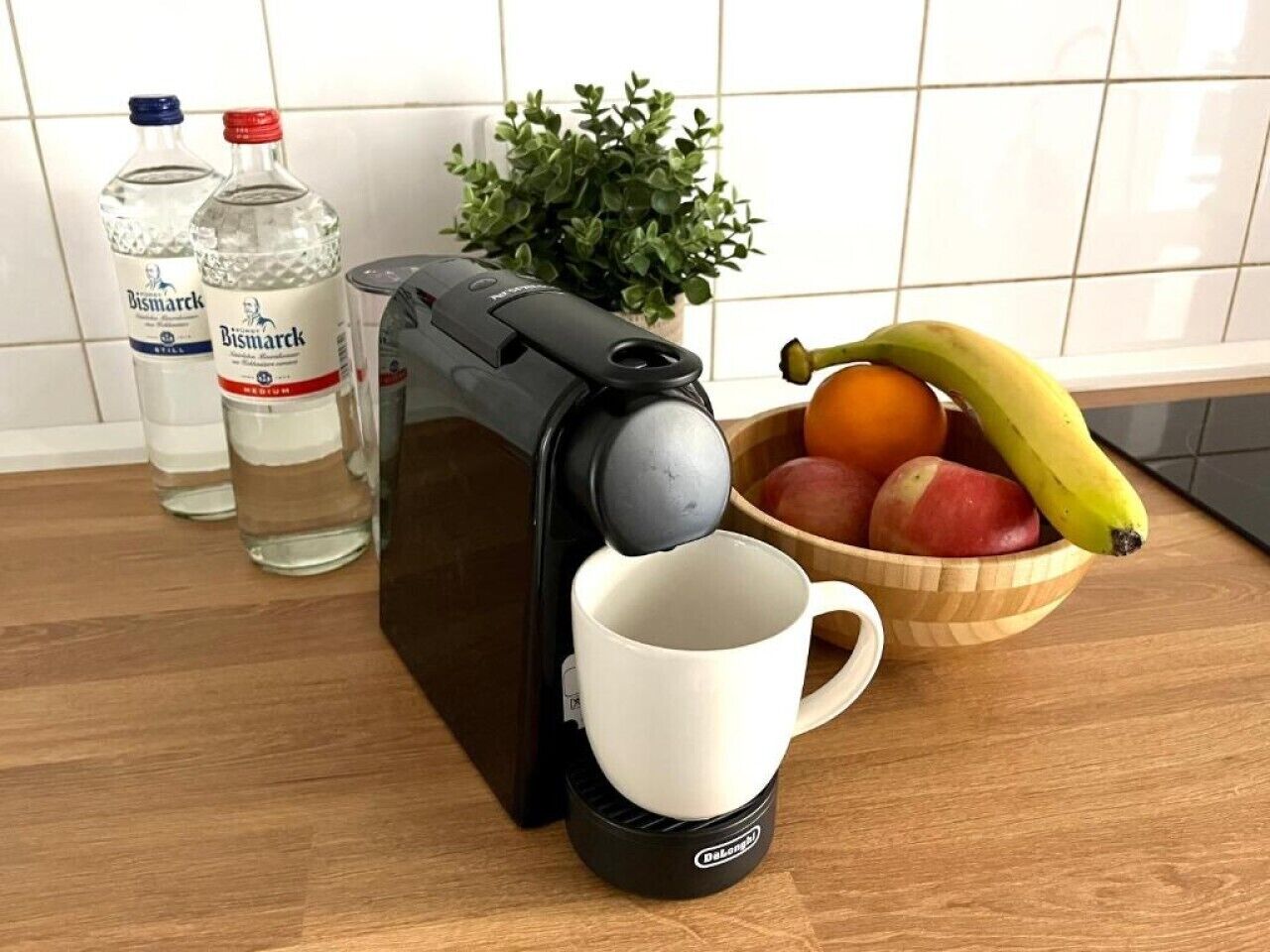 A coffee machine with a cup placed underneath, a bowl of fruit, and two bottles of water on a wooden surface.