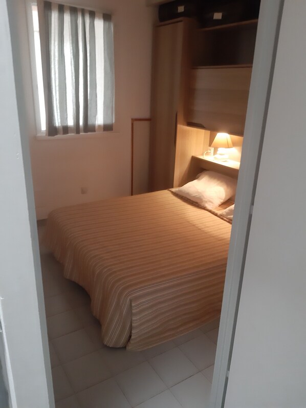 Een kleine kamer met een enkel bed, nachtkastje en lamp, een raam met gordijnen, en een deur die naar een andere kamer leidt.