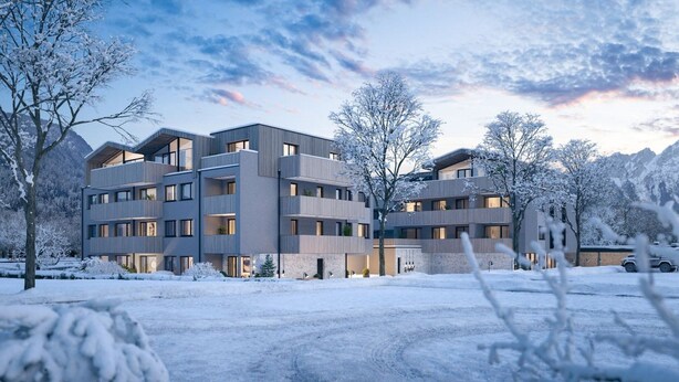 Um edifício moderno de vários andares com varandas, rodeado por árvores cobertas de neve e montanhas ao fundo.