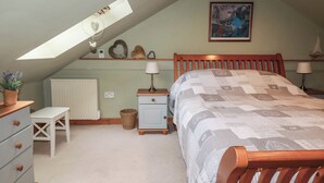 Ein Schlafzimmer mit geneigter Decke, einem Bett mit gesteppter Decke, einem Nachttisch mit Lampe, einer Kommode mit Schubladen, einem kleinen Tisch mit Pflanze und einem gerahmten Bild an der Wand.