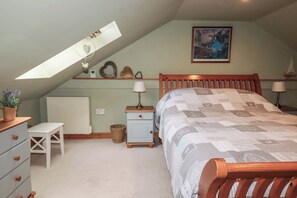 A bedroom with a sloped ceiling, a bed with a patterned quilt, a nightstand with a lamp, a dresser with drawers, a small table with a plant, and a framed picture on the wall. - Skylark (Liskeard)