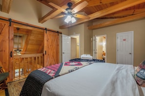 Ein Schlafzimmer mit einem Bett, Deckenventilator, Holzbalken, einer Schiebetür und Blick auf einen Flur.