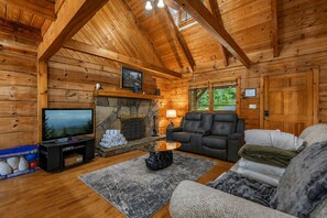 A log cabin interior with a stone fireplace, a television, a sofa, and a large window. - 🏔️ Mountain Views + Hot Tub | Loaded Game Room | Sleeps 10 (Pigeon Forge)