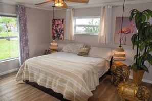 Quarto com cama, duas mesas de cabeceira, ventoinha no teto, janela com cortinas e um vaso de plantas.