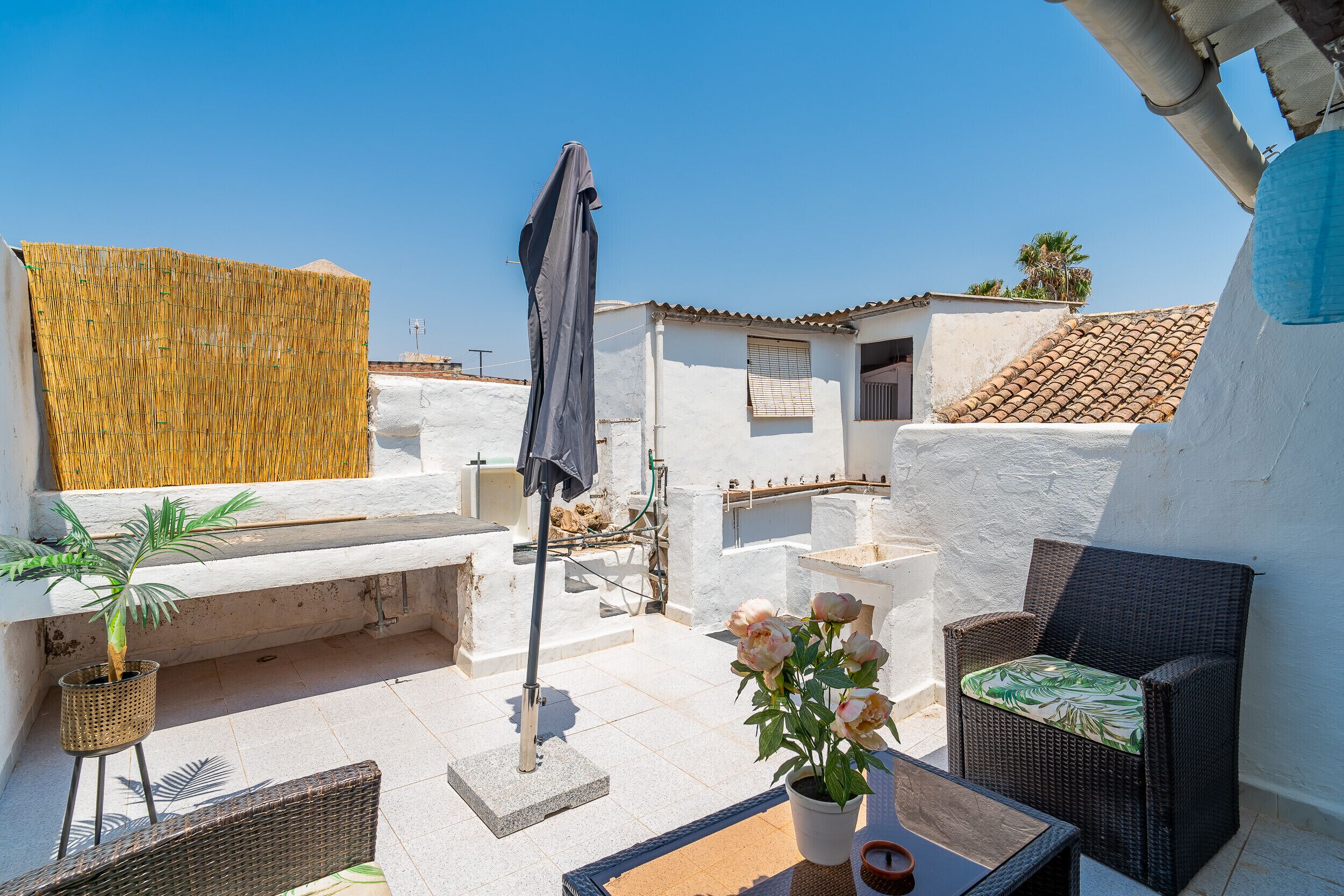 Une terrasse sur le toit avec un mur blanc, une chaise en osier, une plante en pot et une table.