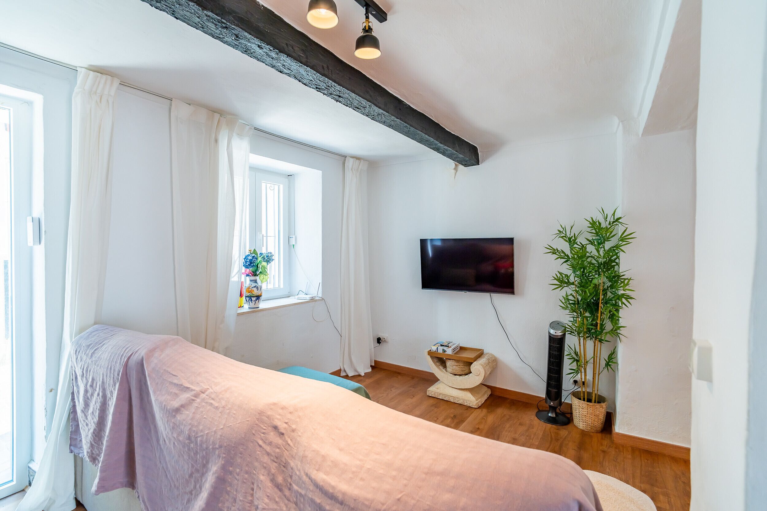 A bedroom with a bed, a window with curtains, a television, a plant, and a wall-mounted air conditioner.