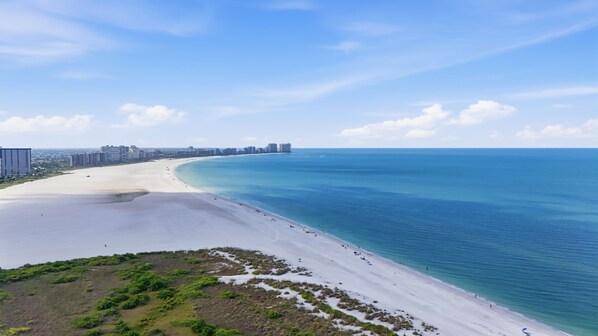 Ein Küstenstrand mit sandigem Ufer, einem klaren blauen Meer und einer Stadtansicht im Hintergrund.