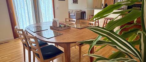 A dining area with a wooden table and chairs, a kitchen in the background, and a large potted plant.