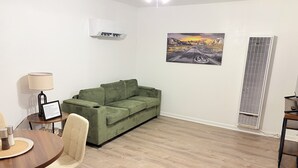 A room with a green sofa, a dining table with chairs, a lamp, an air conditioning unit, a window with blinds, and a painting on the wall. - Route 66 Retreat (Vega)
