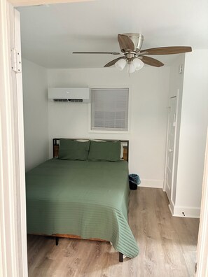 A bedroom with a bed, a ceiling fan, an air conditioning unit, a window with blinds, and a door.