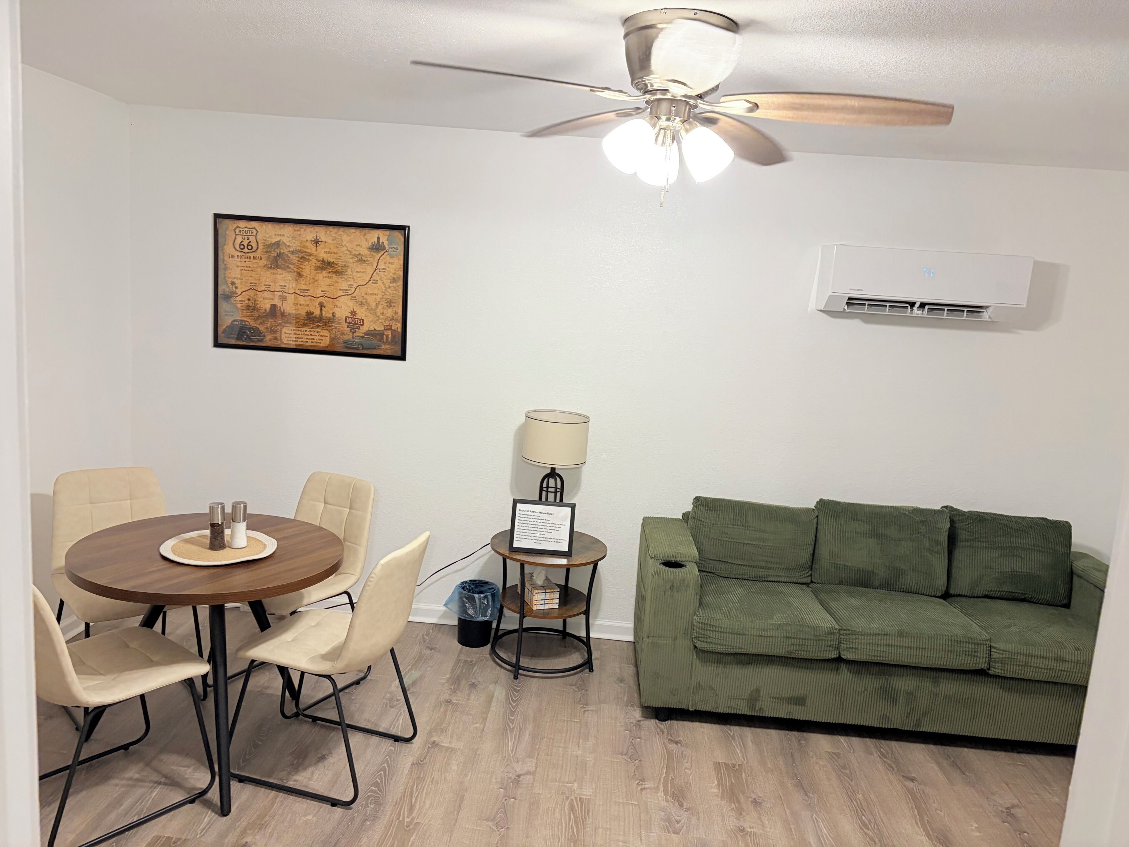 Une pièce comprenant une table à manger, des chaises, un canapé vert, un ventilateur de plafond, un climatiseur mural et une carte encadrée.