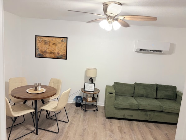 A room with a dining table, chairs, a green sofa, a ceiling fan, a wall-mounted air conditioner, and a framed map.