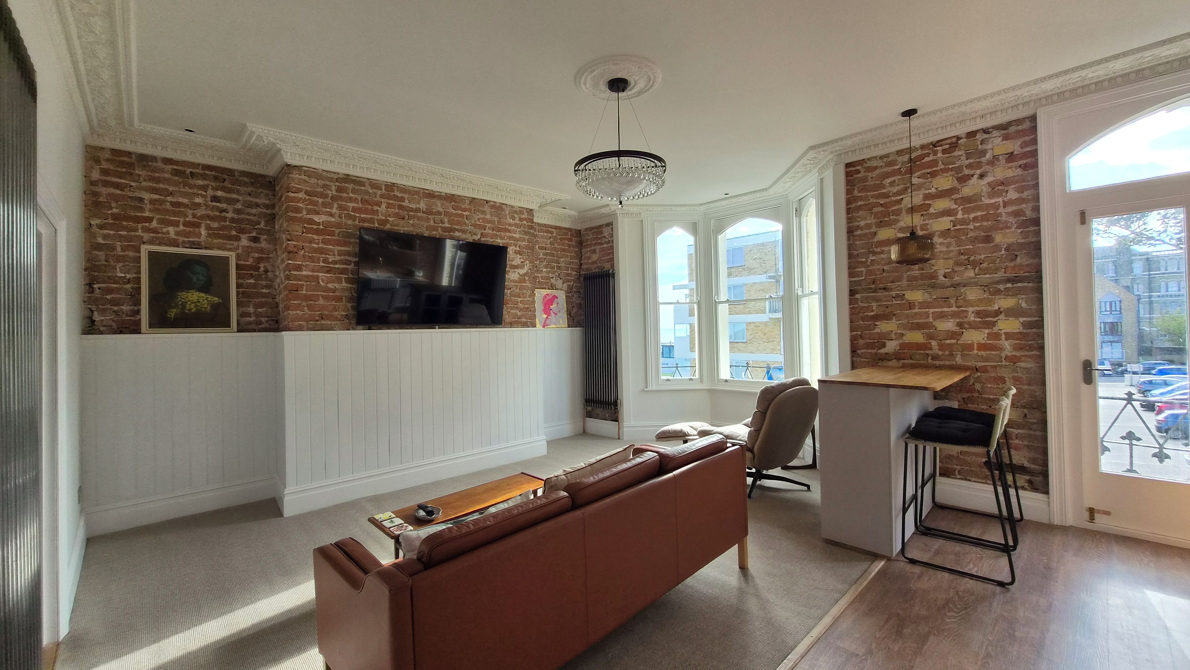 A living room with a brick wall, white paneling, a brown leather sofa, a TV, and a chandelier.
