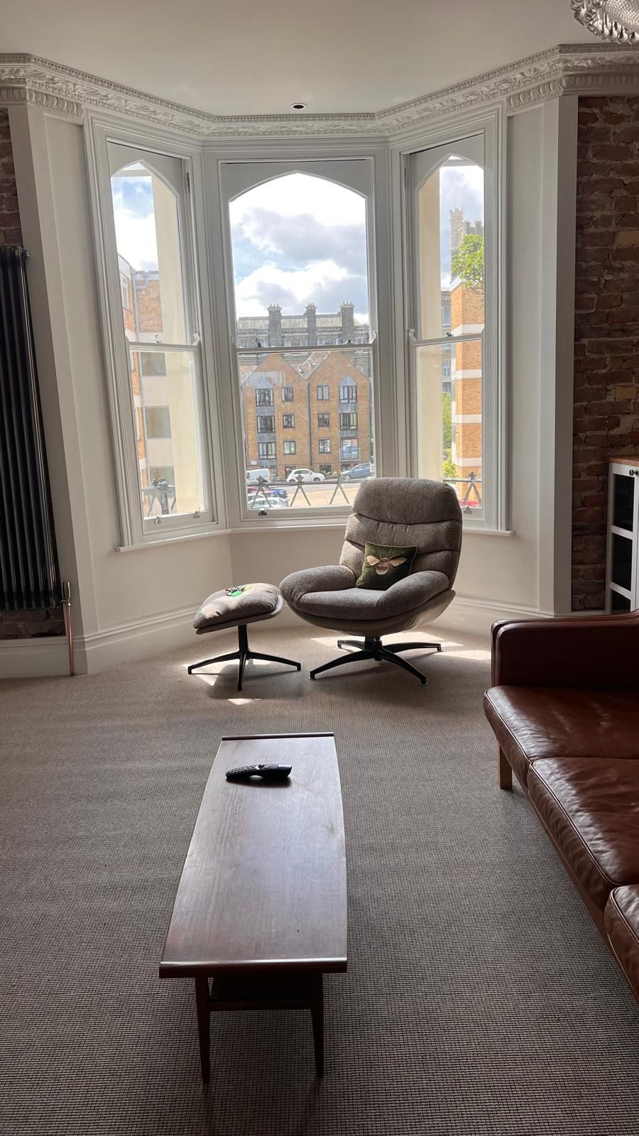 A room with a large bay window, a brown leather sofa, a grey armchair, and a small table.
