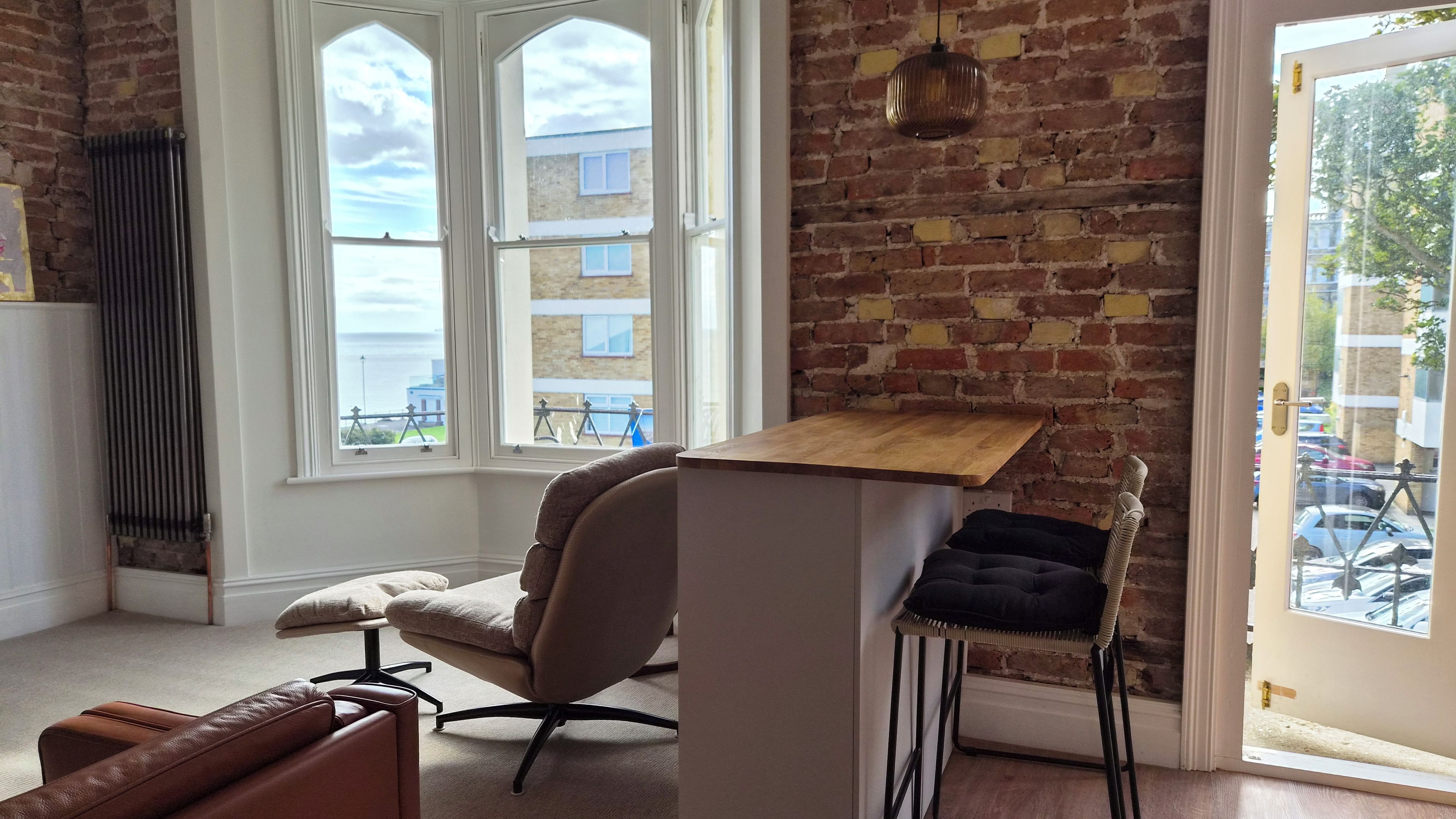 A room with a brick wall, a wooden desk, a cushioned chair, and a view through a window.