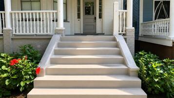 A front view of a house with a set of beige steps leading to a white door, flanked by white railings and green plants.