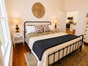 Une chambre à coucher comprenant un lit, des tables de chevet, une lampe, un élément décoratif mural et une vue sur la cuisine.